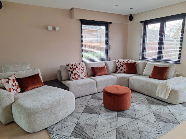 Binnenkijker met ruime lichte hoekbank, fauteuil en roestkleurige woonaccessoires